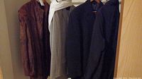 Four men's coats hanging side by side on a rack; brown fur coat, beige plaid coat, dark blue coat, reddish-brown coat
