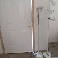 Photo of two white floor lamps side by side near a door. One lamp has a flexible head, the other three lamp heads on a vertical pole.