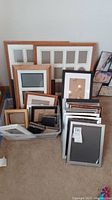 View of multiple assorted picture frames standing and lying against a wall and on the floor, showing a variety of sizes and materials including wood and metal.