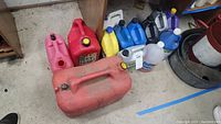 Red and pink empty gas cans along with various automotive fluid containers mostly blue, yellow, purple, and clear plastic jugs seen on floor near wooden shelf.