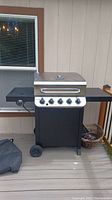 Char-Broil propane grill shown without cover, positioned on a deck. Grill has stainless steel lid with thermometer, black body, five control knobs, and side shelves with one side featuring a side burner.