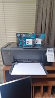 Front angle showing HP DeskJet F4480 printer with paper loaded and two HP 901 ink cartridges on top in original packaging.