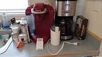 Red Keurig coffee maker, white coffee grinder with power cord, Oster stainless steel drip coffee maker with glass carafe, box of coffee filters on countertop