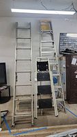 Shows three aluminum ladders leaning against a wall, including one folding step ladder with black steps in front, one 8ft A-frame ladder with yellow top behind, and one extension ladder.