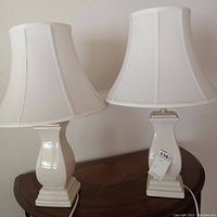 Pair of matching white ceramic table lamps with fabric bell shades shown side-by-side on wooden surface.