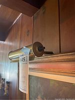 Side view of brass wall-mounted art display lamp above framed artwork on wood paneling.
