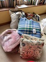 Photo showing wool throws in blue plaid and floral embroidery, pillows with floral embroidery, stacked near windowsill and books.