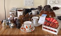 View including glazed ceramic bean pot filled with pine cones, six animal napkin rings, assorted coffee mugs including black, white, yellow, and patterned, two tea cups with saucers having floral and bird designs, and a shrimp-themed napkin holder.