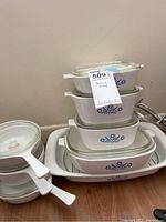 Stack of CorningWare Blue Cornflower casserole and baking dishes with glass lids on wood floor.