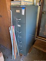 Full body view of the tall 4 drawer metal file cabinet with a few white and orange rods leaning on it. Cabinet is grayish-green and located indoors in a wooden setting.