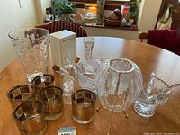 Various glassware items on wooden table including decorated rocks glasses, several vases, bowl and decorative bottle.