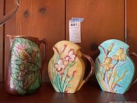 Front view of all three majolica pottery pitchers showing different colors and relief designs.