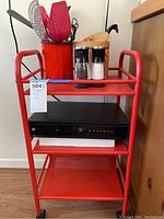 Front and angled views of red metal cart showing tubular rails, three perforated shelves, and wheels.