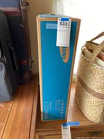 Photo of the sealed Samsung monitor box standing upright on wood floor next to a wicker basket and luggage.