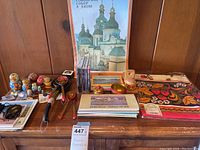 Full view of the Russian memorabilia lot on a wooden table including nesting dolls, decorative eggs, books, pins, salt and pepper shakers in holder, and tablecloth folded in original packaging.