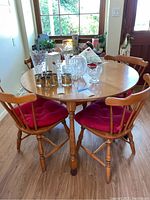 Dining table set with four wooden chairs and red cushions, showing table surface and chairs around it, with various glassware on table.