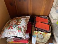 Lot of linens and pillow on curved wooden backing with label visible. Shows embroidered pillow, box with red cloth napkins or placemats, possibly other linens and a few small dishes.