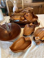 Photo showing large monkey pod wood bowl containing two wooden salad serving utensils, multiple small leaf-shaped bowls, a round wood salad bowl with nutcracker tools, and wooden trays arranged on a table.