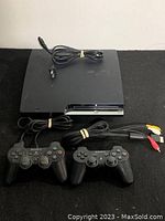 Sony PlayStation 3 slim console with power cable on top, two black DualShock 3 controllers with cables in front, shown on black surface.