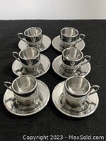Six stainless steel espresso cups placed on their individual matching saucers arranged on a black surface. Cups and saucers have a polished reflective finish and appear in good condition.
