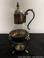 Front side view of vintage glass carafe with ornate silver-plated handle, collar, and lid, placed on silver-plated warmer stand with detailed feet and round platform for heat source.