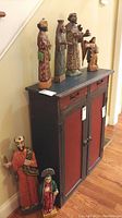 Six carved religious figurines displayed on and next to a black and red cabinet with 2 doors, showing scale and arrangement