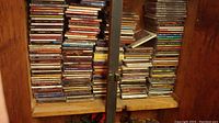 Photo showing stacks of jewel case CDs on a wooden shelf with multiple rows and mixed titles.