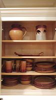 Cabinet shelf displays multiple pieces of handmade pottery including a large pitcher, bear candle lantern, rectangular dish, mugs, bowls, and stacked plates.