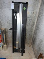 Full view of black mat cutter with metal rail and cutting arm positioned vertically against wall.