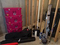 Photo showing ski poles, pink plastic bins, black bag, white helmet, boots, skis leaning against wall