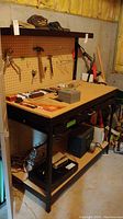 Front angle view of work bench showing metal frame, pegboard, tools on pegboard, wooden top surface, and lower metal shelves with various items (excluded).
