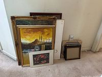 Photo of stacked framed and unframed paintings and multiple panels leaning against wall, showing size and frame styles