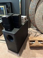 Large black Acoustimass subwoofer speaker with four Bose cube speakers stacked on top and cardboard box on the floor containing wall mounts.