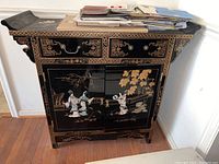 Front view of black lacquer buffet with raised appliqué figures and gold designs, showing drawers and cabinet doors.