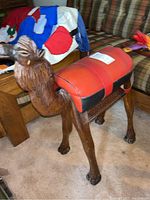 Full side view of carved wooden camel with padded red and black leather seat, placed on carpet next to wooden furniture.
