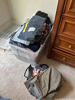 Plastic storage bin filled with folded men's clothing including pants, shorts, and shirts, located next to a wooden dresser.