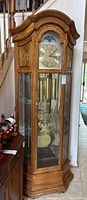 Front view of oak Howard Miller Model 610-440 grandfather clock showing arched bonnet, glass door, mirrored back and glass shelves