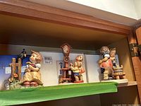 Three Hummel figurines displayed on a shelf with original boxes behind.