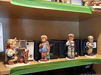 Four Hummel figurines on shelf with their original black boxes behind them