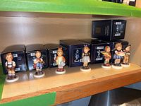 Wide shelf view showing seven Hummel figurines lined up in front of their original black boxes.