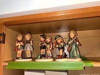 Wide shot of all four Hummel figurines on a wooden shelf showing detailed craftsmanship and colors