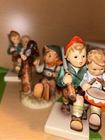 Photo showing two Hummel figurines: one child holding a rifle and another playing drum, standing close together, with a third sitting child figurine blurred behind.