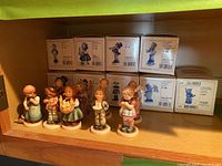 Nine Hummel porcelain figurines displayed on shelf with original boxes in the background.