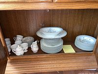 Wide shot of the Noritake Ireland china set displayed in wooden cabinet showing various pieces including cups, cream, sugar and stacked plates.