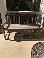 Wooden porch bench photographed outdoors in sunlight, showing front view with slatted back and seat.