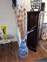 Full view of the tall blue and white ceramic vase with faux flowers and branches inside, placed on hardwood floor beside a dark wooden cabinet.