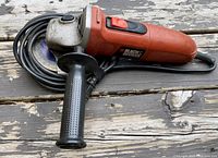 Side perspective showing the angle grinder with side handle, power cord coiled, red and black body, and grinding disc.