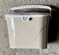 Photo of a beige plastic countertop compost bin with a white handle and lid taken from the front on a concrete surface