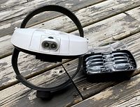 Headset head mounted magnifier with black adjustable headband and dual LED lights on front, placed on wood surface with lens case next to it.