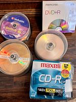 Photo showing multiple spindles and packs of blank writable discs including Verbatim DVD+R DL 15-pack, Memorex DVD+R 5-pack, two unbranded spindles, and sealed Maxell CD-R 12-pack.